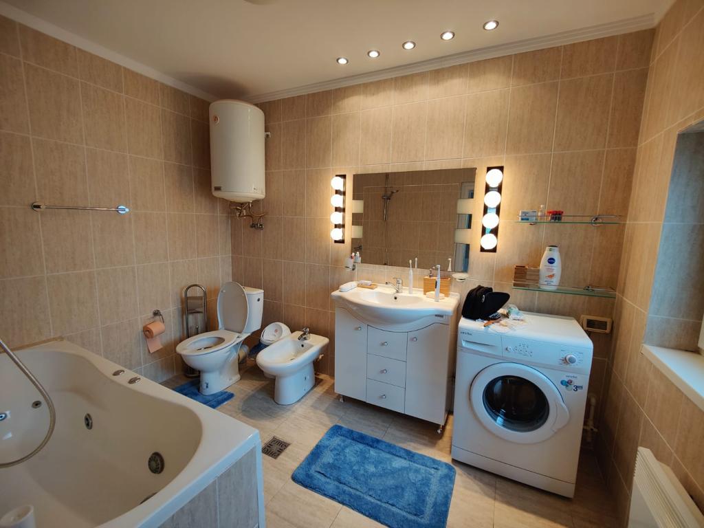 The spacious bathroom features warm-toned tiles and modern fixtures. A large whirlpool tub sits adjacent to two toilets. A double vanity with a large mirror is illuminated by vanity lights. A washing machine is positioned against one wall, adding to the room's functionality.