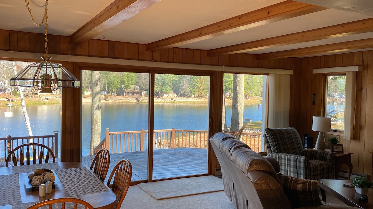 A comfortable living area is presented, featuring a large window that offers a view of the serene lake. Wooden paneling enhances the warmth of the space, complemented by a dining table set for six. A cozy sofa is positioned near a coffee table, inviting relaxation.