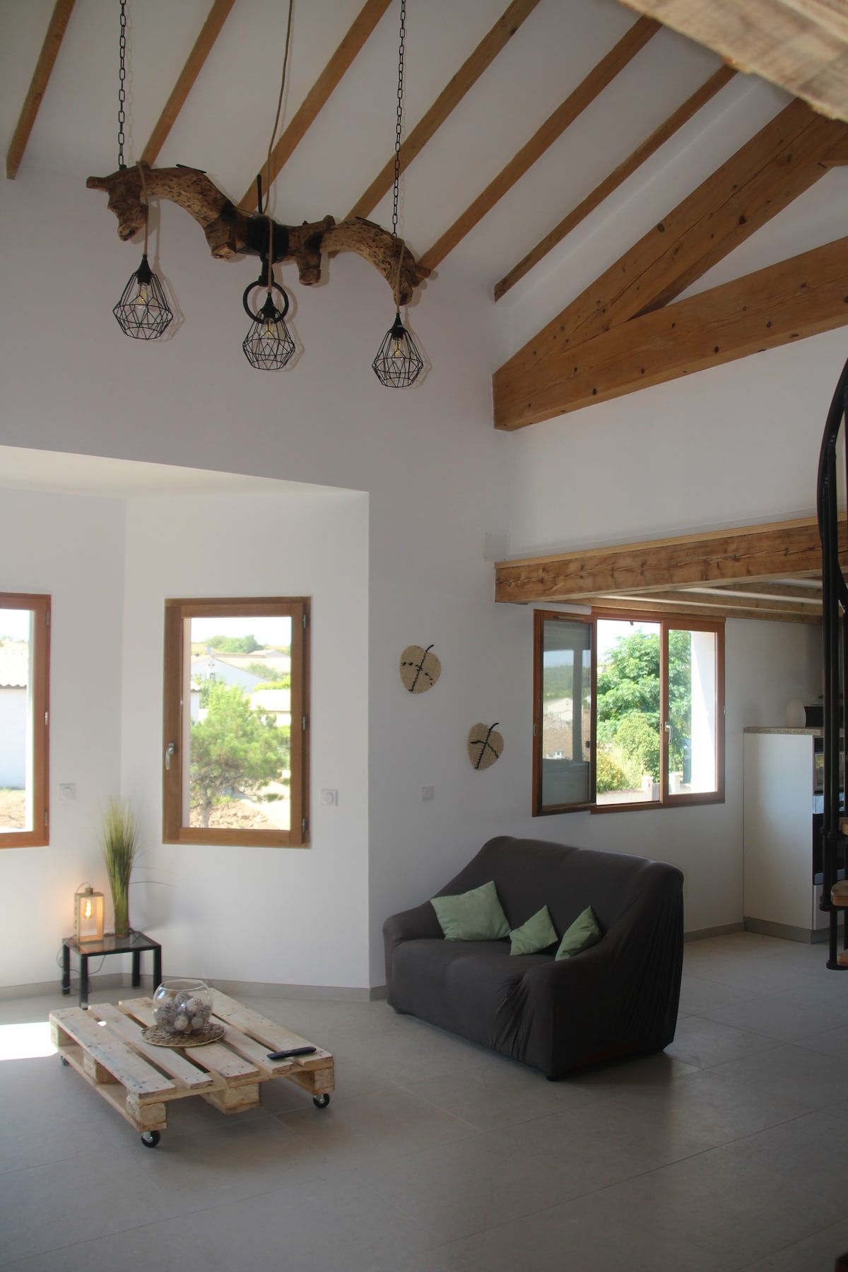 A bright living space features high ceilings with exposed wooden beams and distinctive pendant lighting. Large windows provide natural light, while a gray sofa offers comfortable seating. A low coffee table made of wooden pallets adds a rustic touch, complemented by simple decor and greenery.
