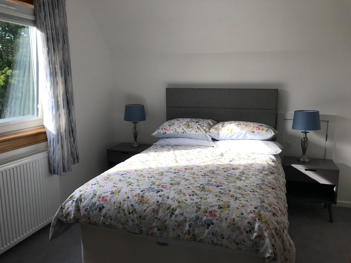 A bright double bedroom features a spacious bed topped with a floral-patterned bedding set. Bedside tables with blue lamps are positioned on either side. Natural light streams in through a window, complemented by light curtains that softly frame the view.