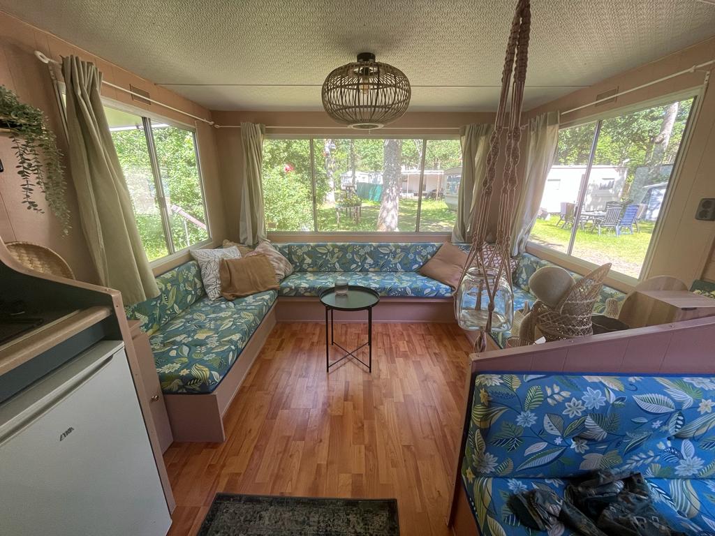 A bright and airy living space features large windows, allowing ample natural light. Comfortable seating arrangements with floral-patterned cushions are situated around a small coffee table. A decorative hanging light and a cozy rug enhance the welcoming feel of the room.