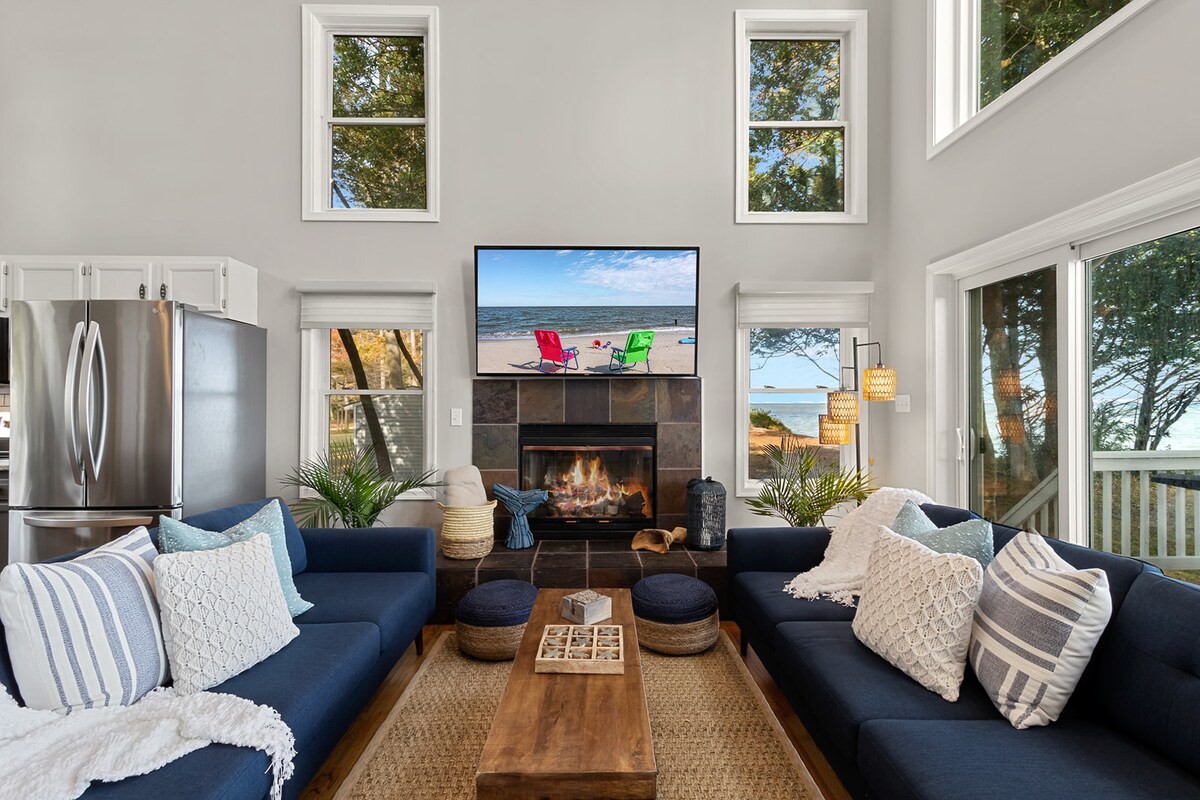 A modern living area features two dark blue sofas adorned with various textured cushions, arranged around a wooden coffee table. A large flat-screen TV is mounted above a sleek fireplace, while windows on three sides allow natural light and views of the surrounding trees and beach.