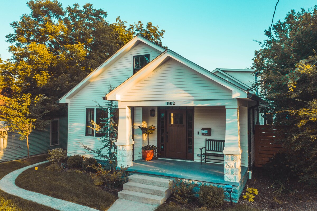 The exterior of a charming house is shown, featuring a welcoming front porch with a bench and planters. Lush greenery surrounds the home, and a curved walkway leads to the entrance. Large windows allow natural light to highlight the inviting facade.