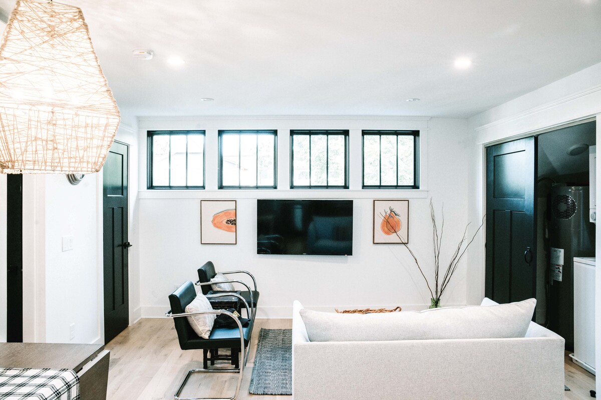 A light-filled living space features a cozy sofa and sleek armchairs. A mounted television is positioned across from the seating, with large windows providing ample natural light. Artwork is displayed on the walls, adding visual interest to the room.