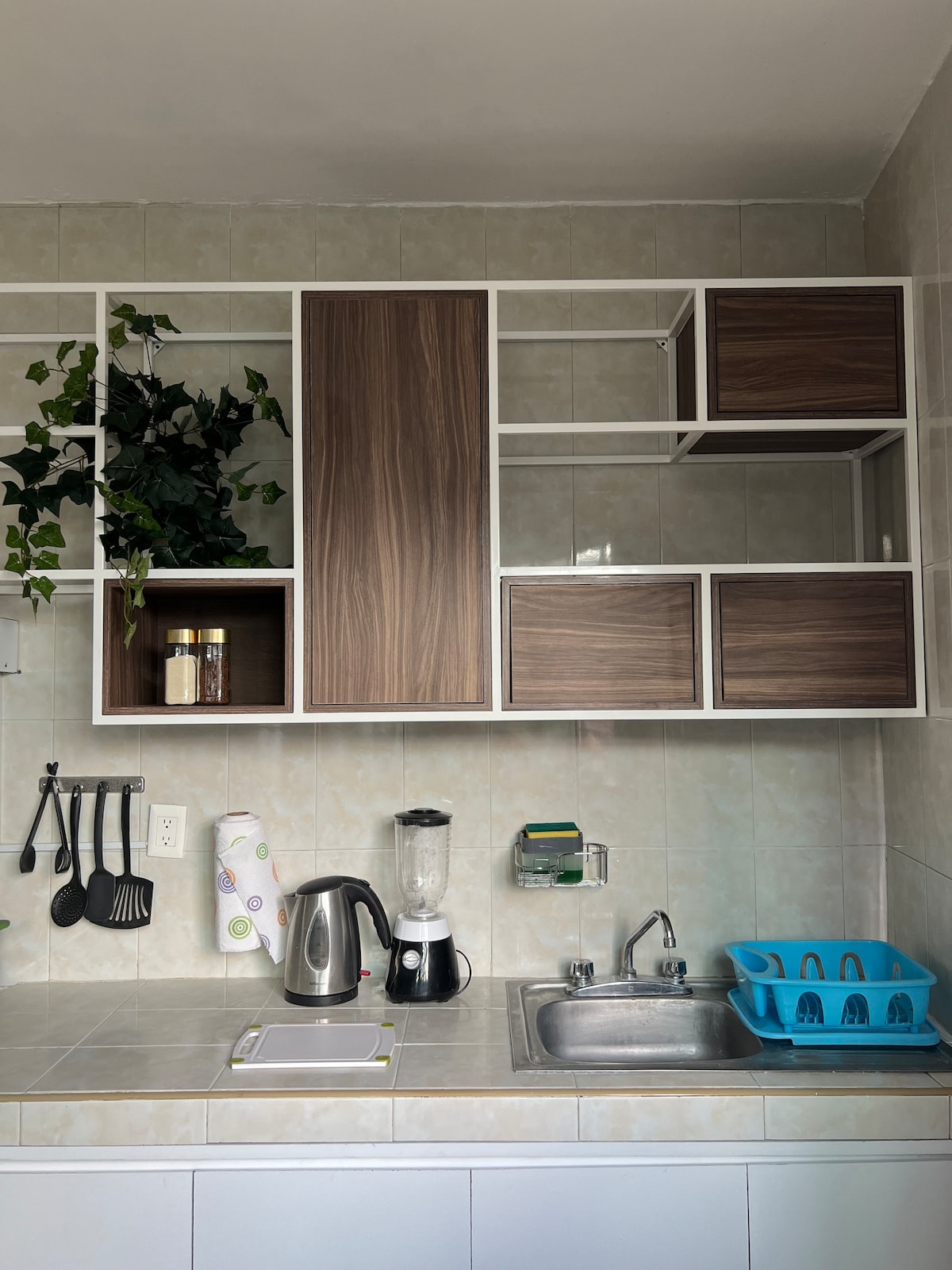 A compact kitchen space features wall-mounted wooden storage units with a combination of open shelving and closed cabinets. A silver kettle, a blender, and various kitchen utensils are visible. A small sink is positioned to the right, alongside a light blue dish rack for drying dishes.