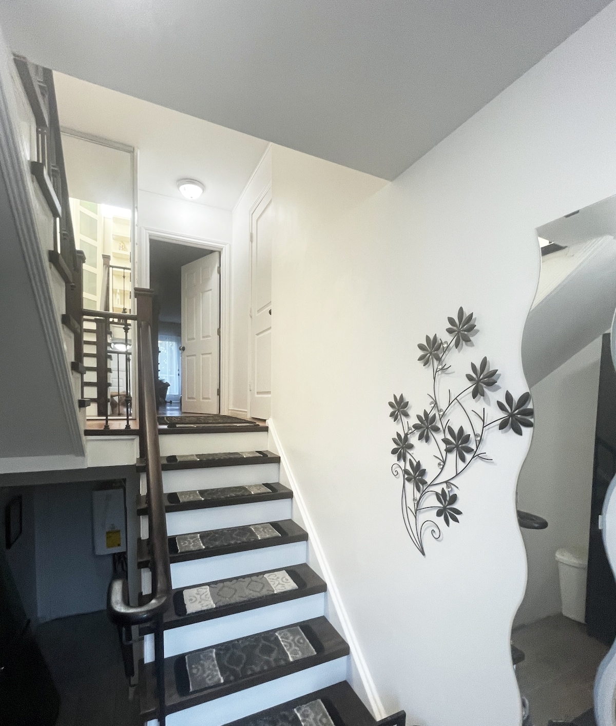 A well-lit staircase features wooden steps with decorative tiles and a floral wall art piece accentuating the light-colored walls. The stairs lead to upper levels, while a mirror reflects the space, enhancing the openness of the area.