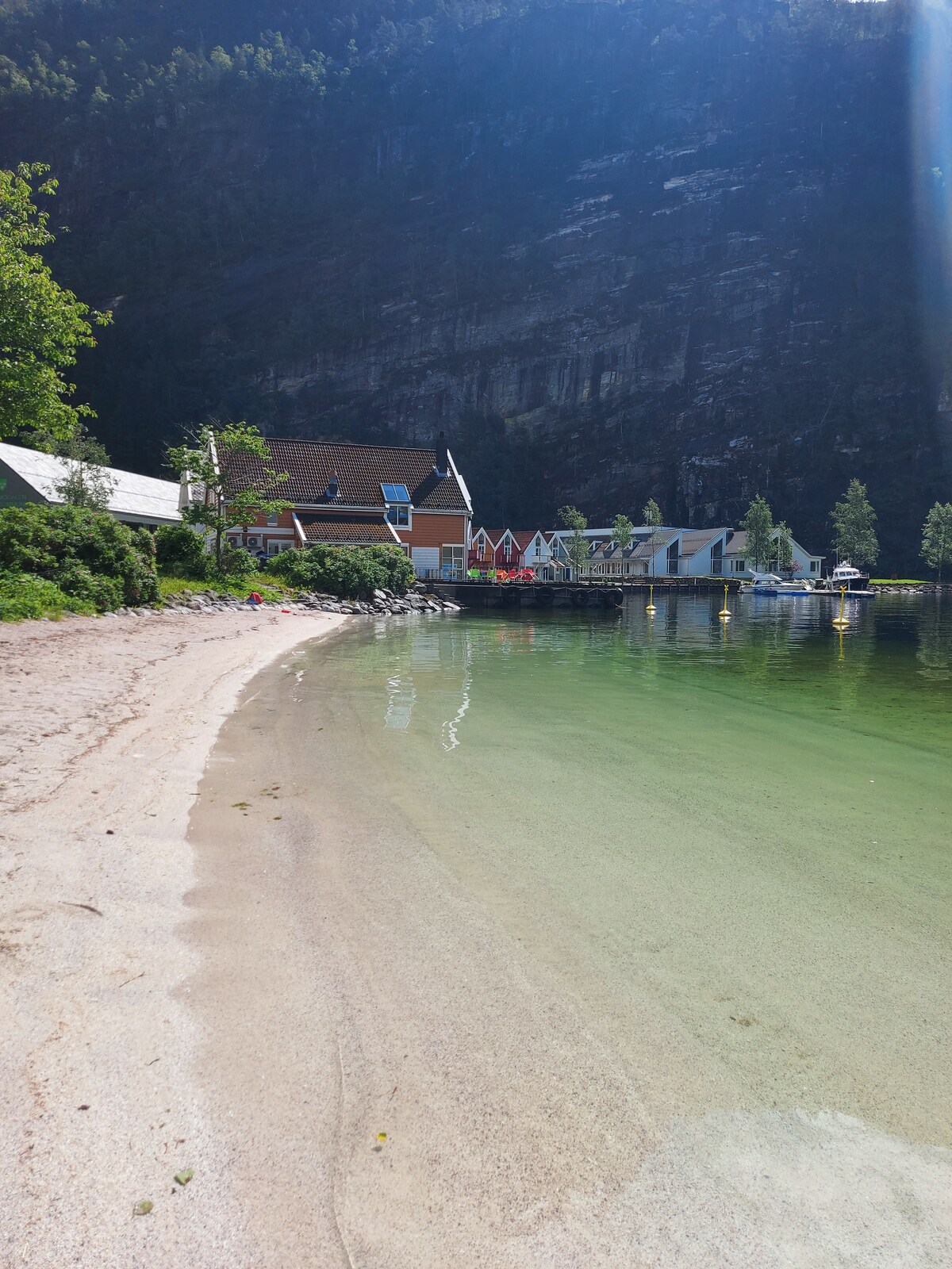 A calm sandy beach is visible along the water's edge, with clear greenish water reflecting the surrounding landscape. Towering cliffs rise in the background, while several buildings are positioned along the shoreline, creating a scenic and serene atmosphere.