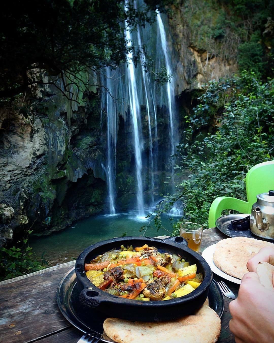 A rustic outdoor dining setup features a steaming dish of colorful vegetables and meat served in a traditional pot. In the background, a stunning waterfall cascades down rocky cliffs, surrounded by lush greenery. The scene combines natural beauty with a sense of culinary delight.