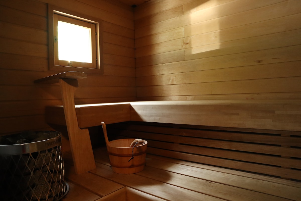 A serene sauna space features wooden paneling throughout, with a cozy bench for seating. A small window allows natural light to enter, and a wooden bucket and a metal basket are present, contributing to the authentic sauna experience.