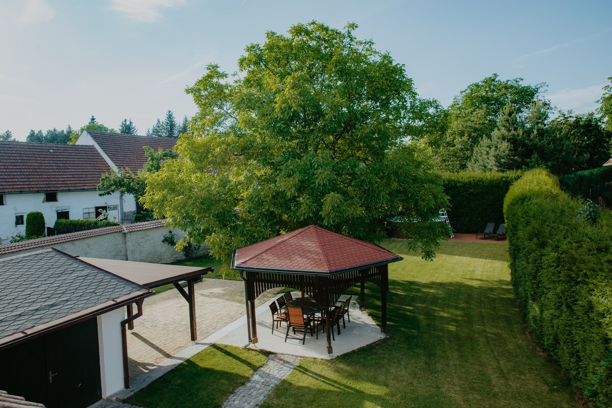 A spacious garden area is highlighted by a large gazebo with a wooden seating arrangement. Lush green grass surrounds the gazebo, while a shaded tree provides additional comfort. A stone path leads to the outdoor space, surrounded by neatly trimmed hedges.
