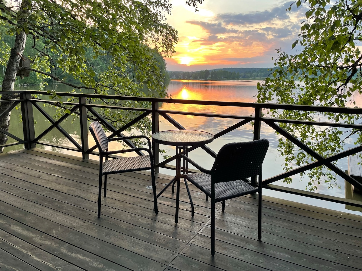 A serene outdoor terrace is depicted, featuring a round table accompanied by two chairs. The scene captures the gentle glow of sunset reflecting on the lake's surface, framed by trees and wooden railing that encourages relaxation amidst nature.