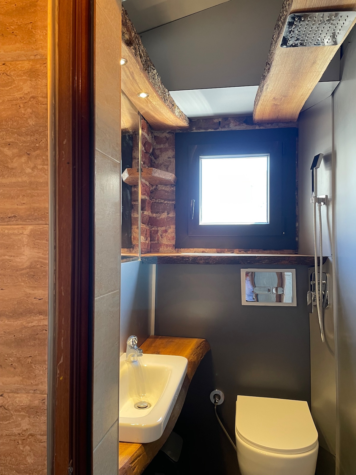 A compact bathroom features a modern sink with a wooden countertop and a toilet set against a textured wall. Natural light enters through a small window above the sink, and a showerhead is mounted on the wall, creating a functional yet stylish space.