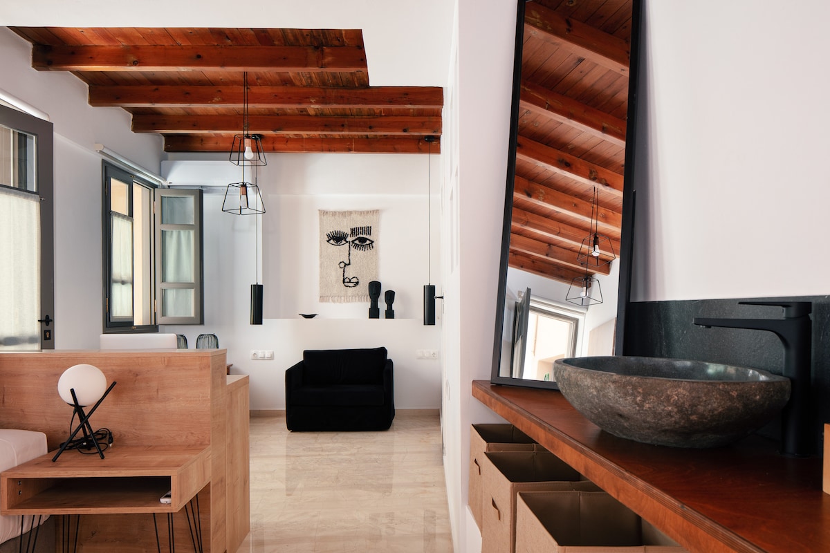 An open-plan living space is highlighted by wooden beams on the ceiling and features large windows allowing natural light. A black sofa adds comfort, while a minimalist art piece and decorative sculptures enhance the modern decor. The stylish sink and wooden cabinetry provide functional elements.