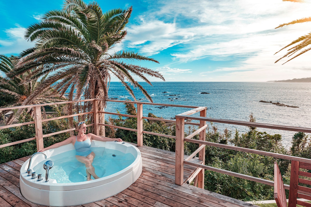 A private jacuzzi is situated on a wooden deck, surrounded by lush palm trees and offering panoramic views of the ocean. The sky features a blend of soft blues and warm hues, while gentle waves can be seen in the distance.
