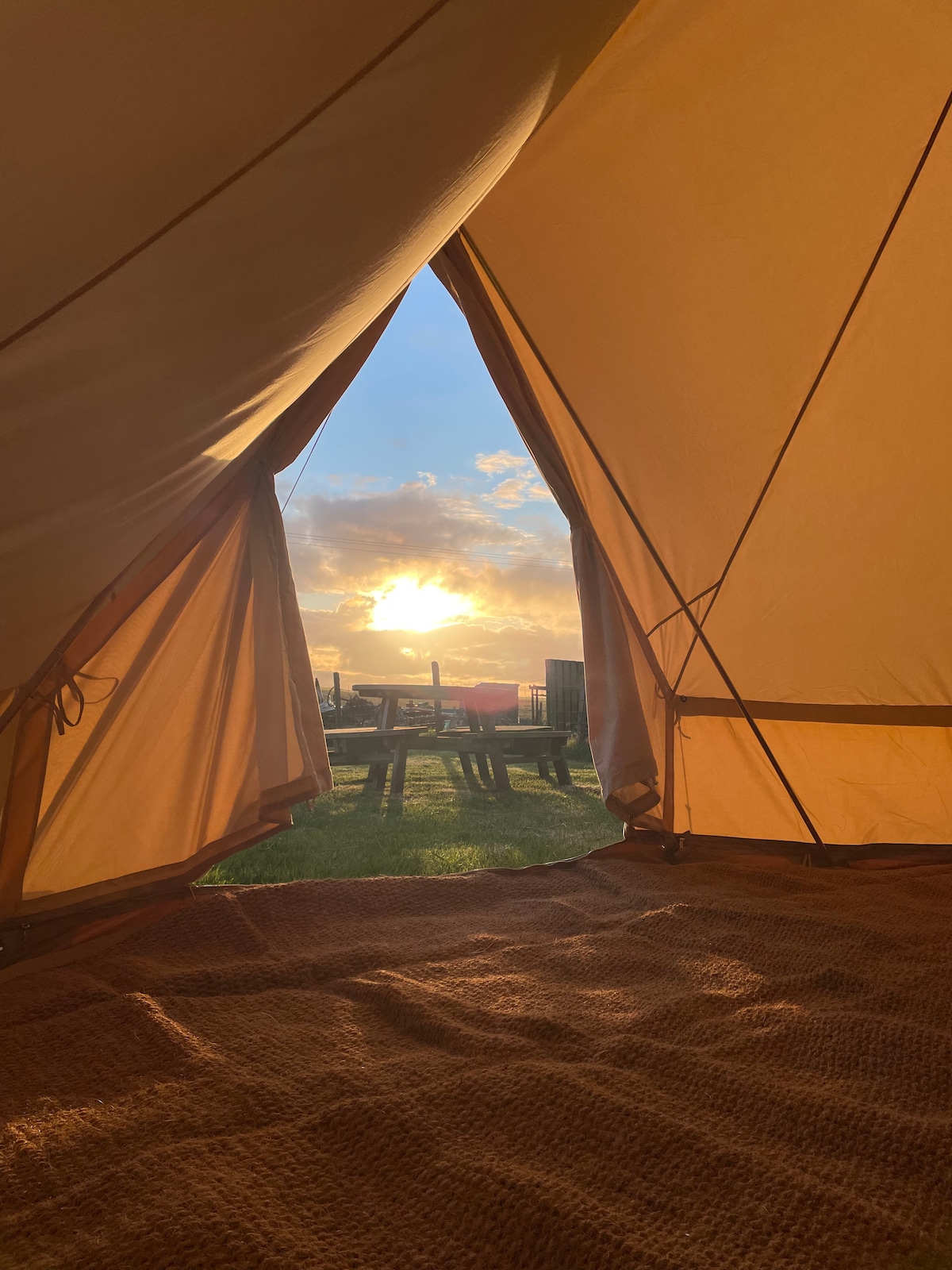A view from inside the canvas bell tent captures a sunrise, with soft golden light filtering through the open entrance. Outside, wooden furniture is visible against a backdrop of lush greenery, creating a serene connection with nature.