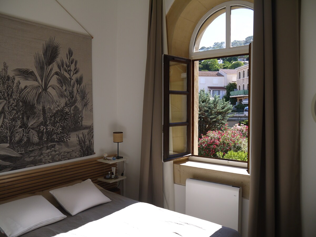 A serene bedroom is depicted with a spacious bed adorned with neutral bedding. Natural light enters through a large arched window, framed by soft curtains. Outside, a glimpse of a flowering garden and nearby buildings can be seen, enhancing the tranquil ambiance.