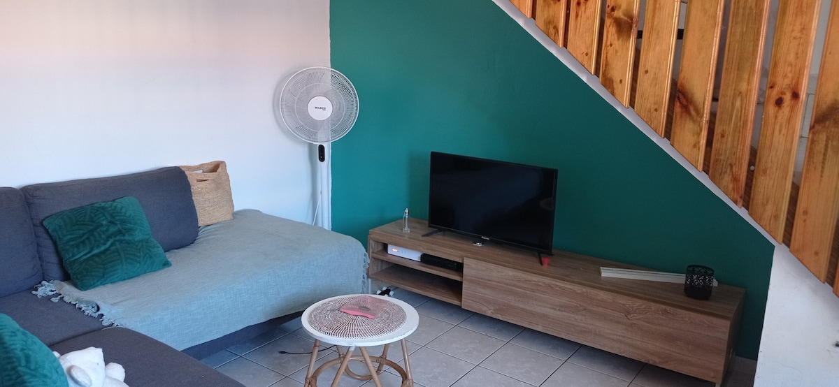 A comfortable living area features a gray sofa and a light blue throw blanket. A wooden television stand holds a flat-screen TV, while a small round table completes the space. A wall-mounted fan is positioned nearby for added comfort.