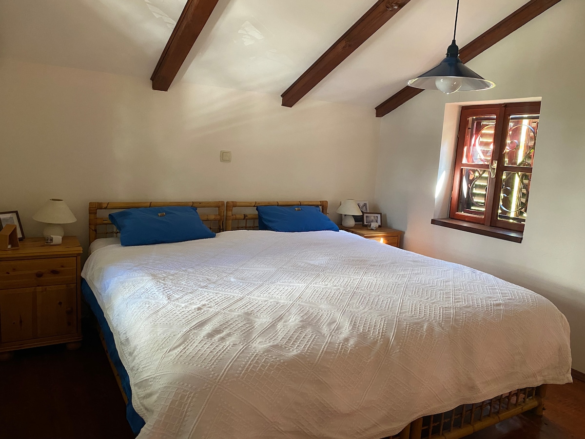 A spacious sleeping area features a large bed with a white quilt and two blue pillows. Natural light filters through a small window adorned with wooden shutters, while two bedside tables are positioned on either side of the bed, each topped with a lamp.