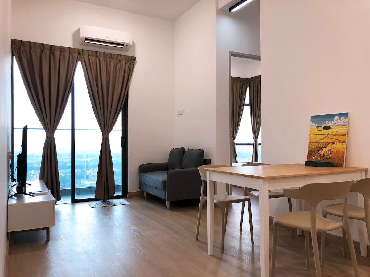 A modern living area features a plush gray sofa and a light wood dining table surrounded by four chairs. Large glass doors offer scenic views, with curtains elegantly framing the windows. A flat-screen television is positioned on a minimalist console.