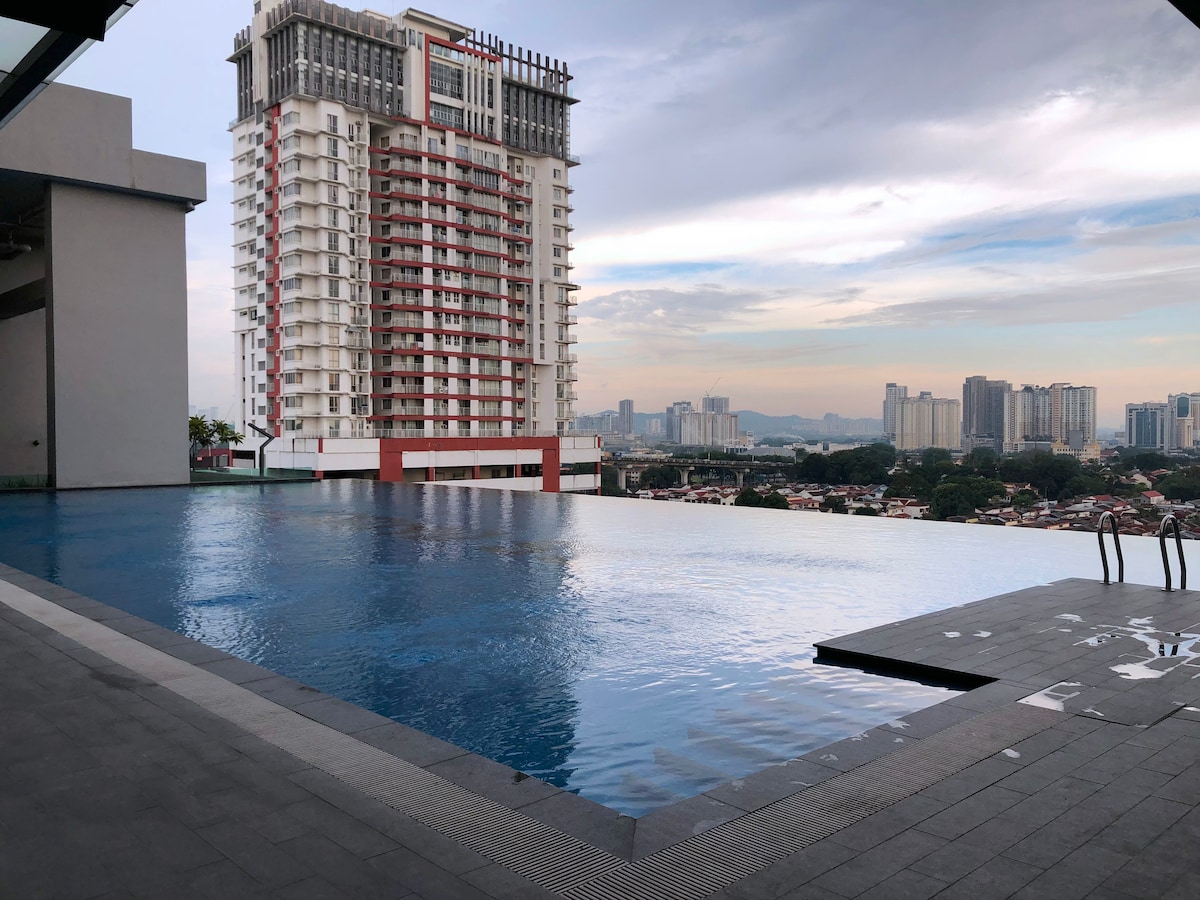 An infinity pool extends along the edge of a terrace, reflecting the surrounding skyline. The clear water merges with the horizon, while modern buildings rise in the background under a softly lit sky.