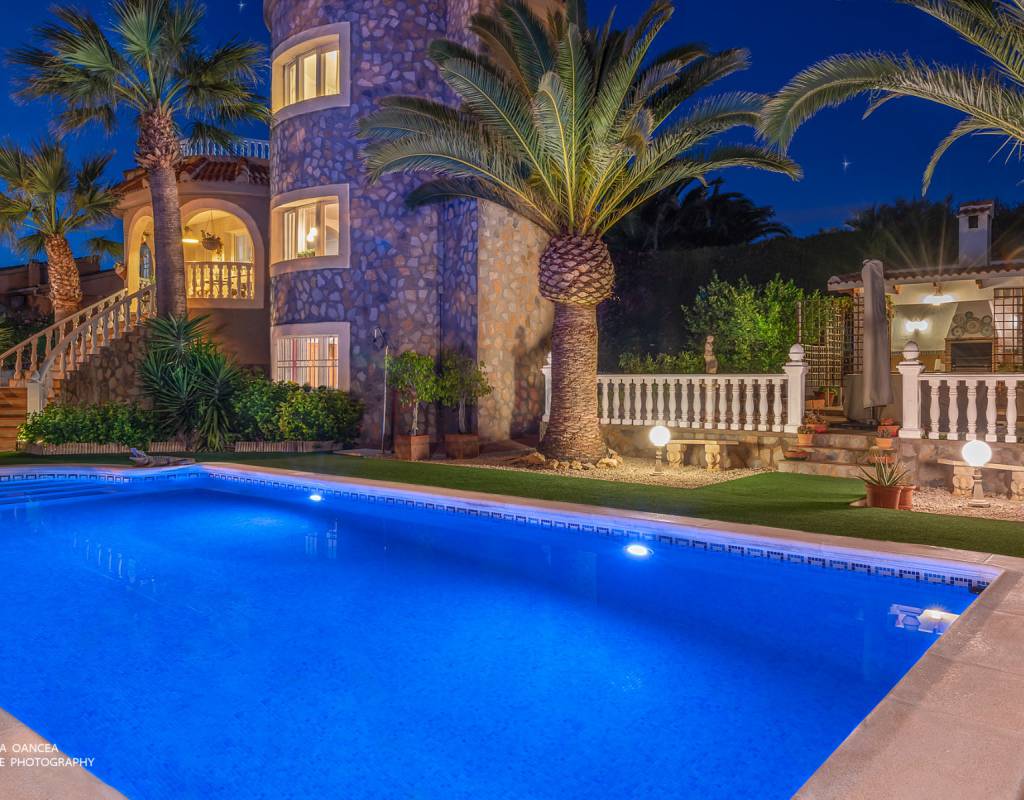 The outdoor area features a vibrant blue swimming pool, surrounded by lush greenery and palm trees. The villa's stone façade and decorative railings create an elegant backdrop, all illuminated by soft lighting at dusk.
