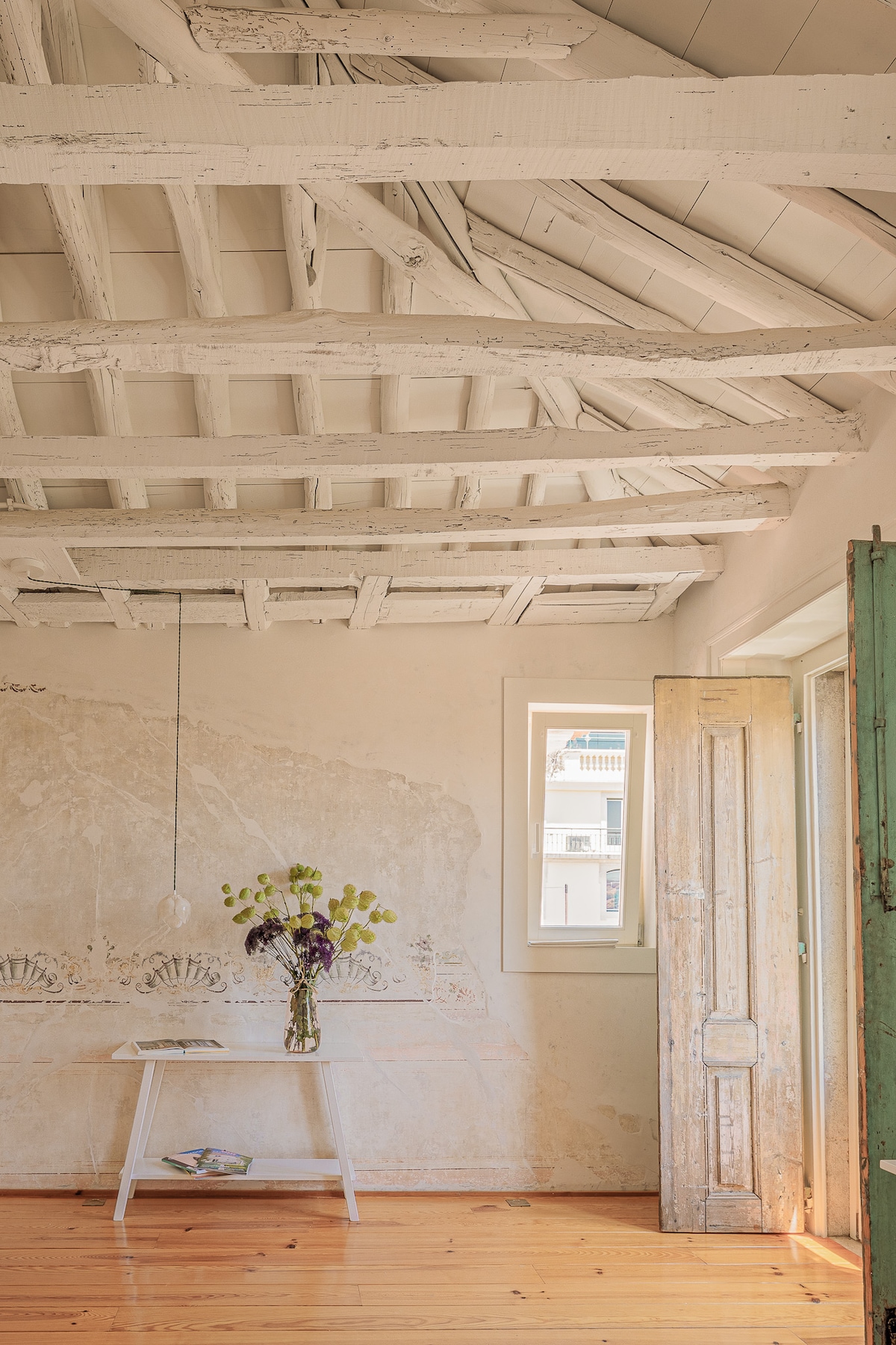 A bright interior space features whitewashed wooden beams and warm wooden flooring. A small white table holds a vase of fresh flowers. Natural light enters through a window beside a partially open door, revealing subtle wall textures and a minimalist aesthetic.