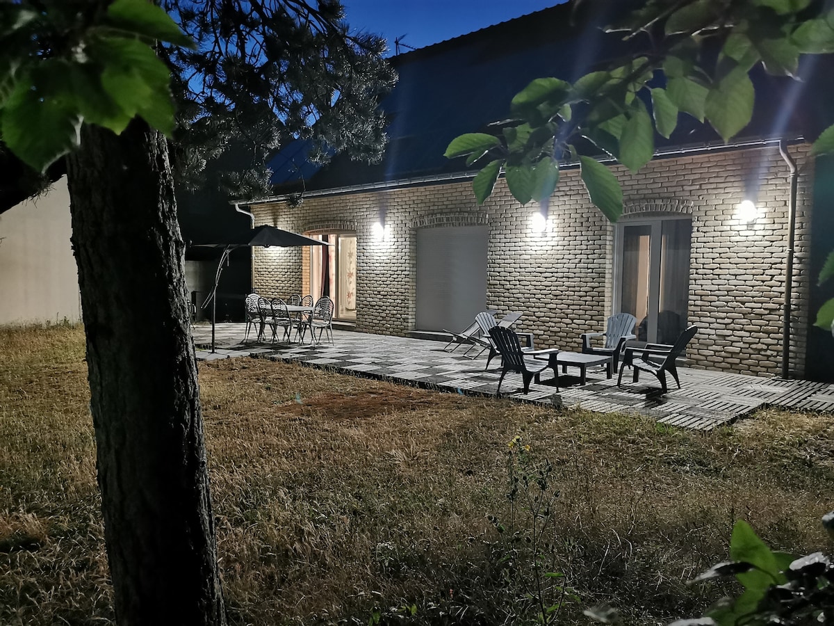 The exterior patio area is softly illuminated in the evening, featuring a seating arrangement with multiple chairs and a table. The surrounding grass is trimmed, and trees frame the space, providing a serene backdrop to the brick façade of the house.