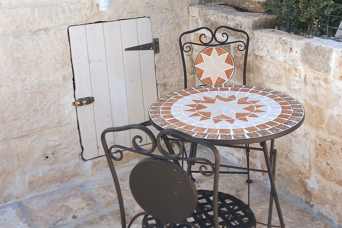 A mosaic-topped bistro table is accompanied by a wrought iron chair, situated in a serene outdoor space. A small door with a latch is set into the stone wall, adding character to the setting.