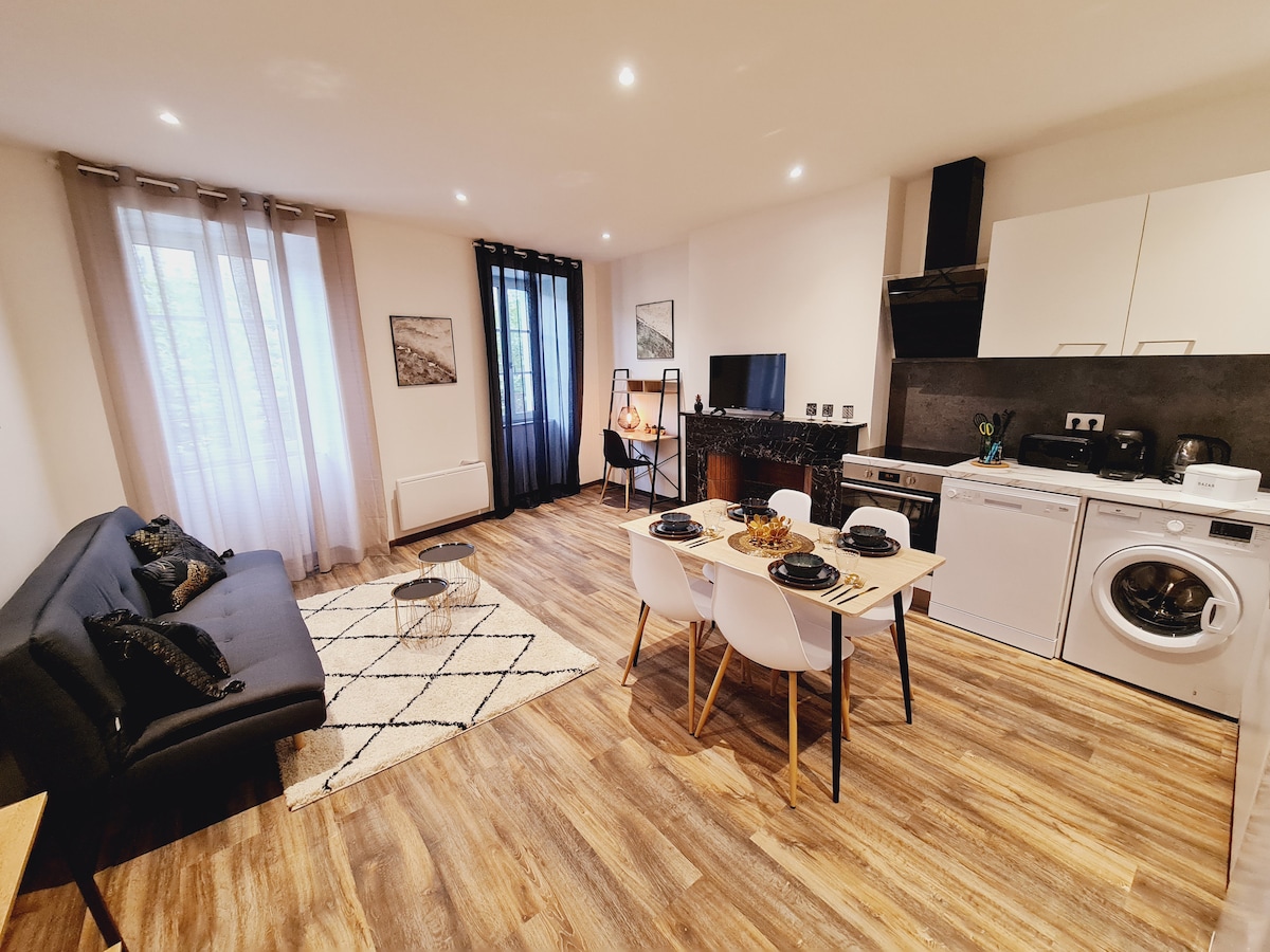 An open-plan living area is displayed, featuring a black sofa facing a wall-mounted television. A dining table with white chairs is set with dinnerware. Light wood flooring complements the modern kitchen appliances, including a washing machine. Natural light enters through large windows, enhanced by sheer curtains.
