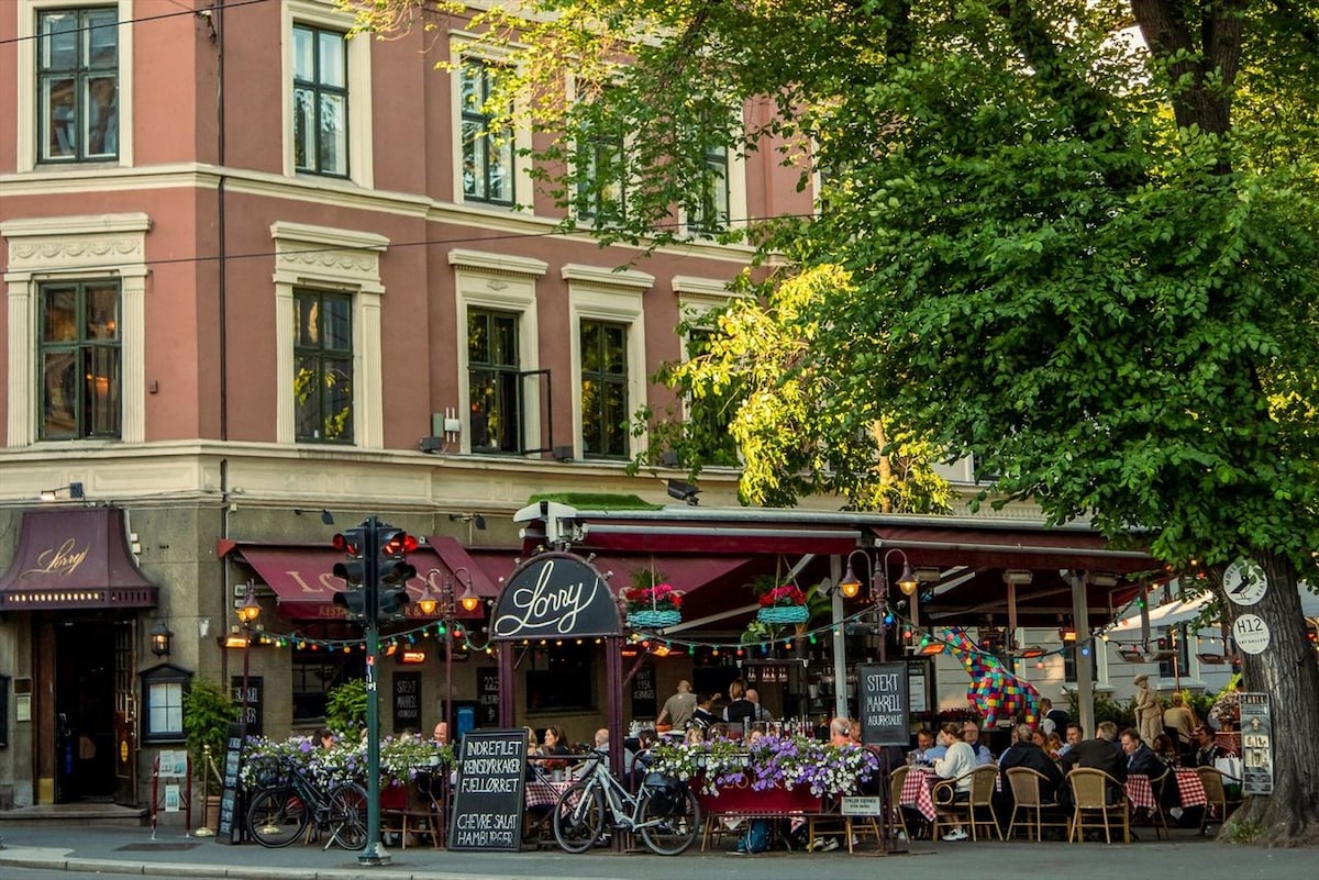 An outdoor dining area is adorned with vibrant flowers and surrounded by green trees. Patrons are seated at tables under an awning, enjoying their meals. A bicycle rack can be seen nearby, and the building's facade features large windows and a classic aesthetic.