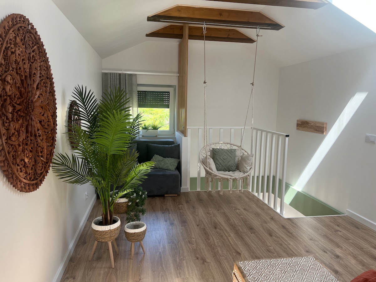 A bright indoor space features a comfortable seating area with a dark sofa and decorative cushions. A swinging chair is suspended from the ceiling, enhancing the room's openness. Potted plants add a touch of greenery, and natural light filters through a nearby window.