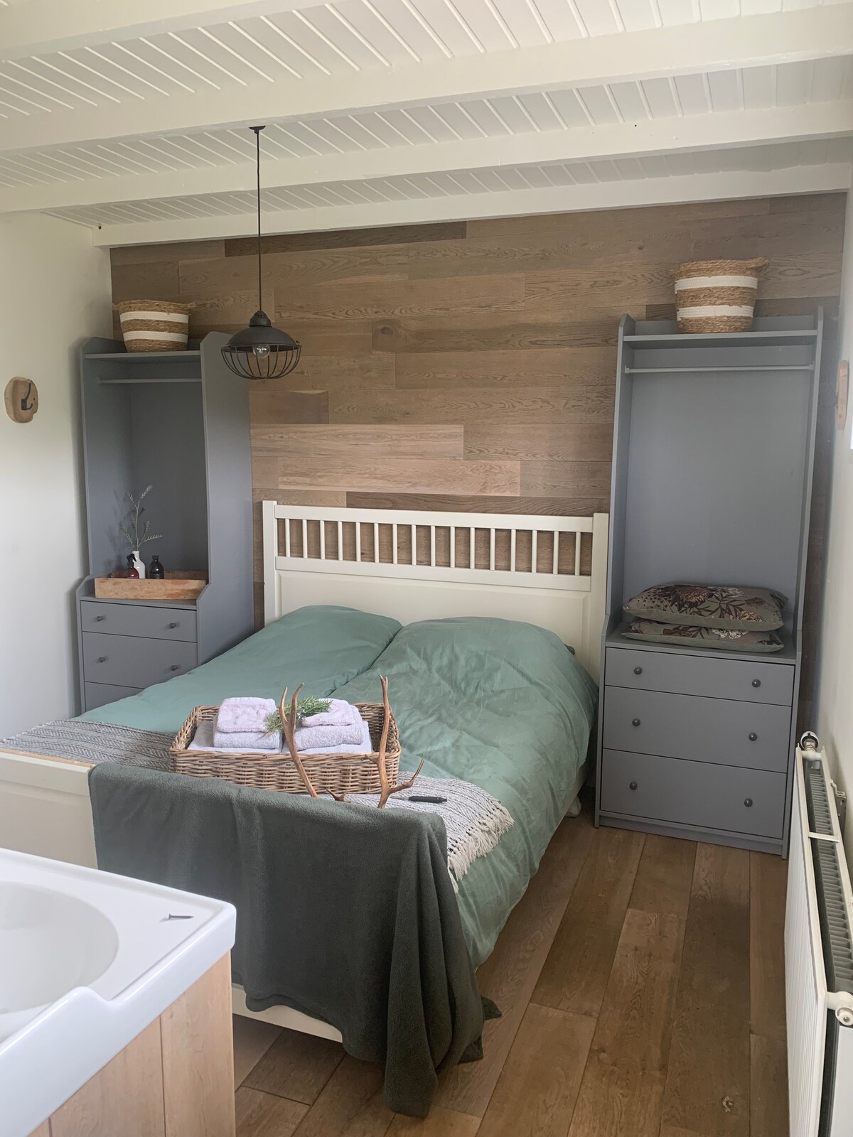 A cozy bedroom features a spacious bed dressed in light green linens. Flanking the bed are two grey cabinets. A woven basket containing towels is placed on a linen-covered surface, complemented by a soft throw draped over the bed. Light wood accents provide a warm ambiance.