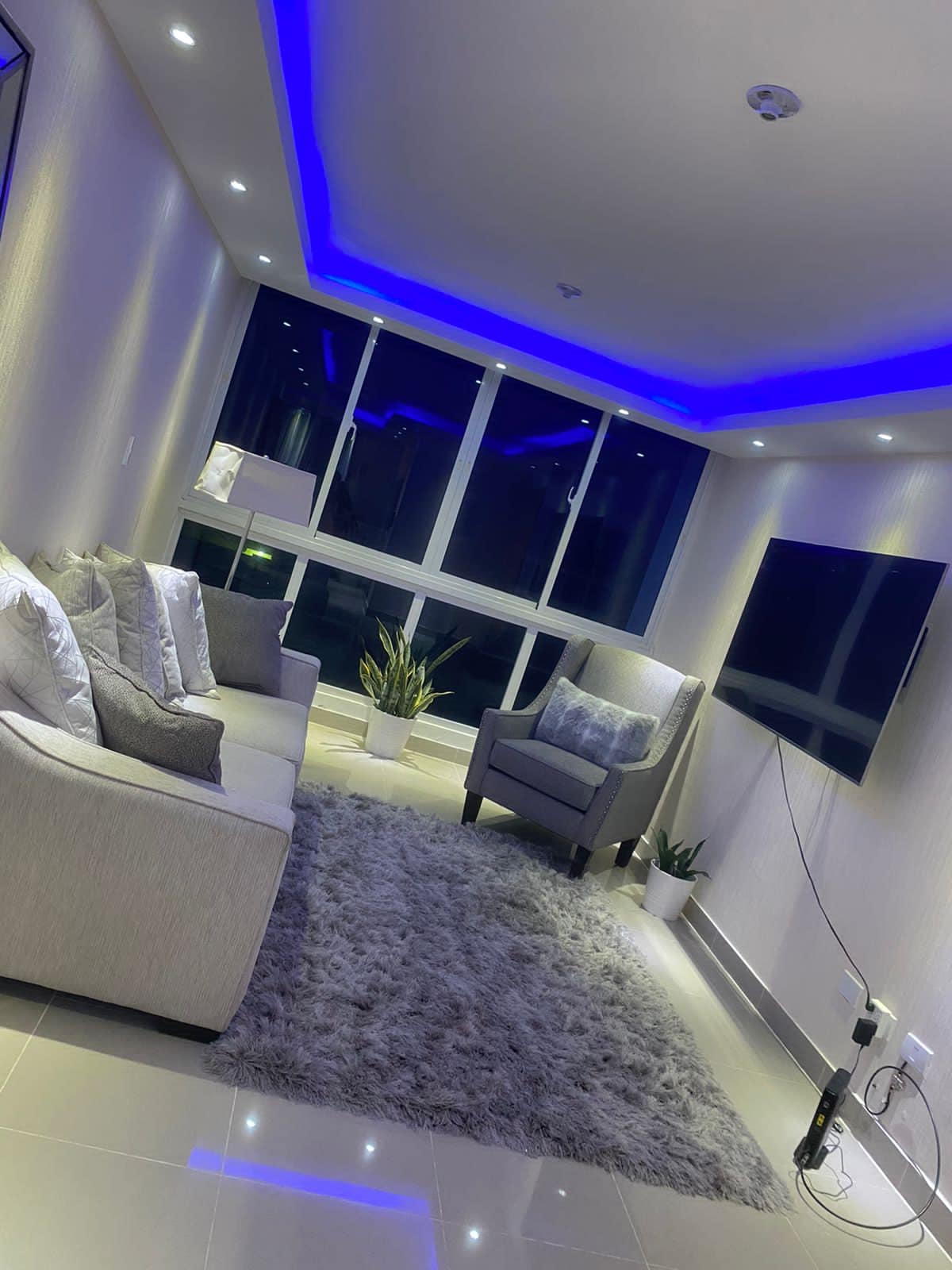 A well-lit living area features a plush gray sofa and an armchair positioned on a soft area rug. Soft blue lighting illuminates the ceiling, and a large window offers a view of the surrounding area. A simple plant adds a touch of greenery.