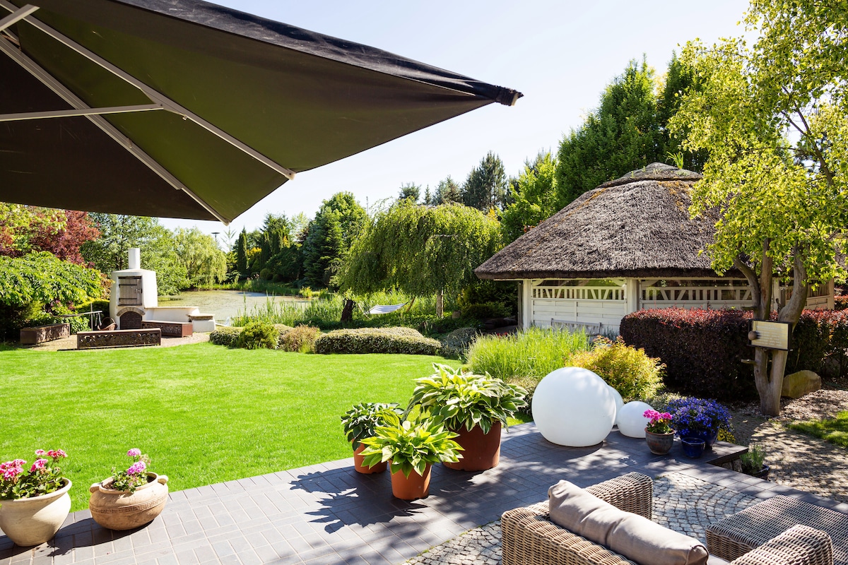 A spacious garden area is featured, showcasing a manicured lawn surrounded by various plants and flowers. A thatched-roof gazebo is visible in the background, complemented by a modern outdoor seating arrangement. A sunshade provides relief from sunlight on this serene outdoor space.