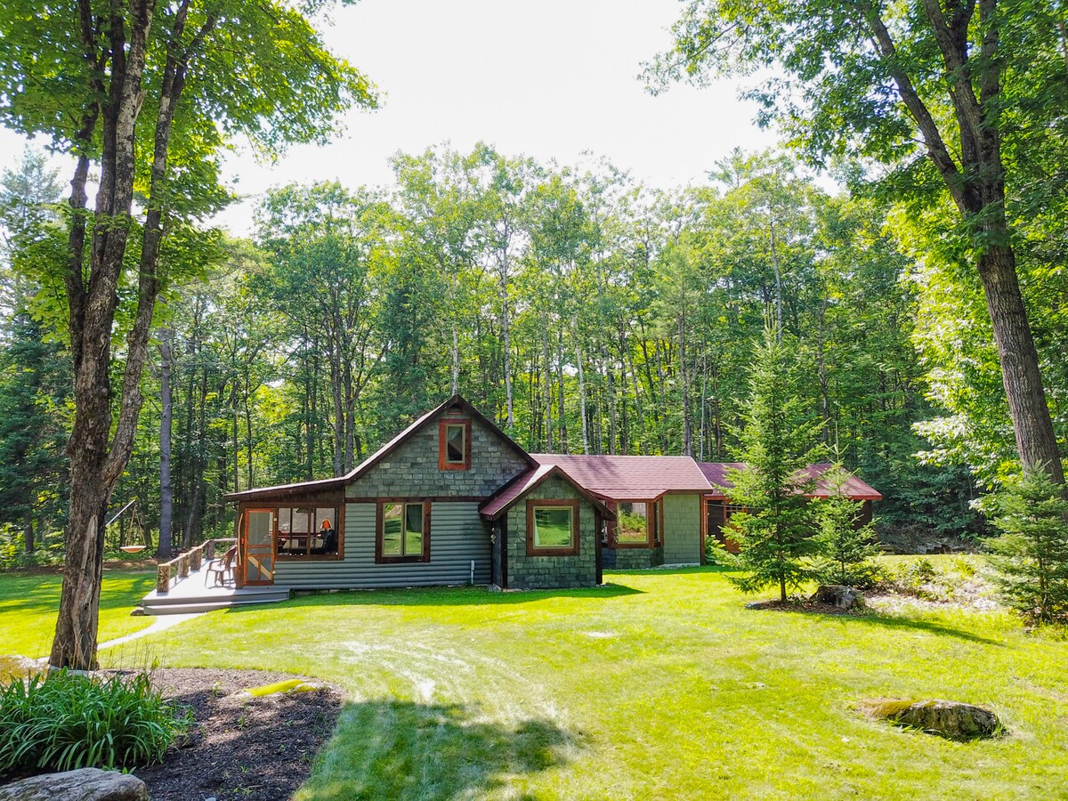 A rustic cabin is nestled within a woodland glade, surrounded by tall trees and a spacious, green lawn. Sunlight filters through the leaves, highlighting the inviting exterior, which features a mix of wooden and textured siding, along with a welcoming front deck.