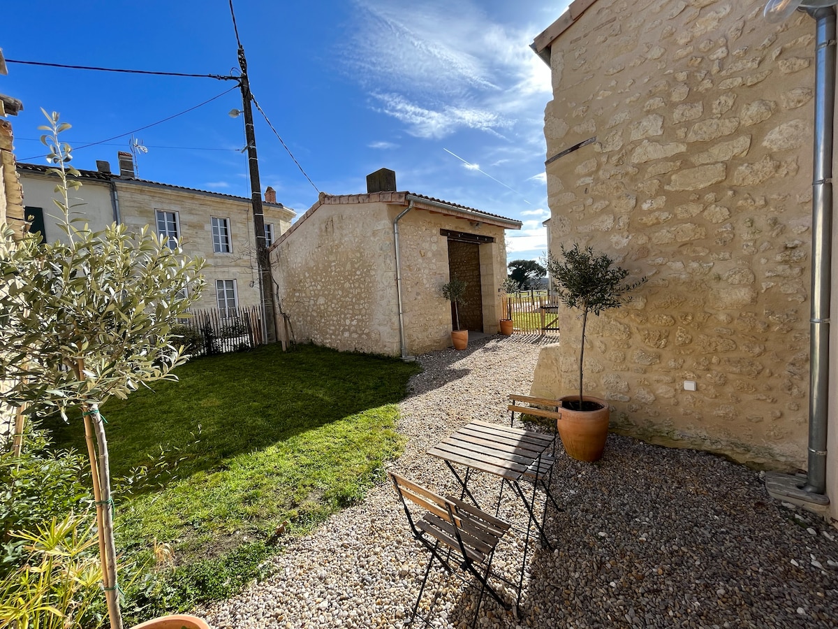 An outdoor area is presented, featuring a small gravel courtyard with a green lawn. Two wooden chairs and a table are arranged beside a terracotta pot. A stone building stands in the background, under a clear blue sky with a few wispy clouds.