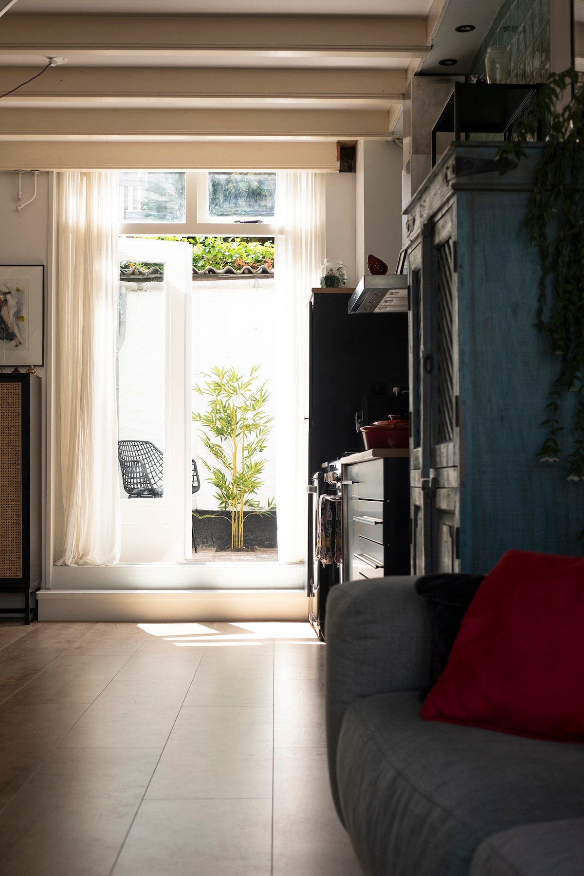 Natural light fills the room, highlighting a view of a small outdoor patio with a leafy plant. The modern kitchen area is visible, featuring sleek black cabinetry and a hint of a stove. Soft textures in the nearby seating area add warmth to the space.