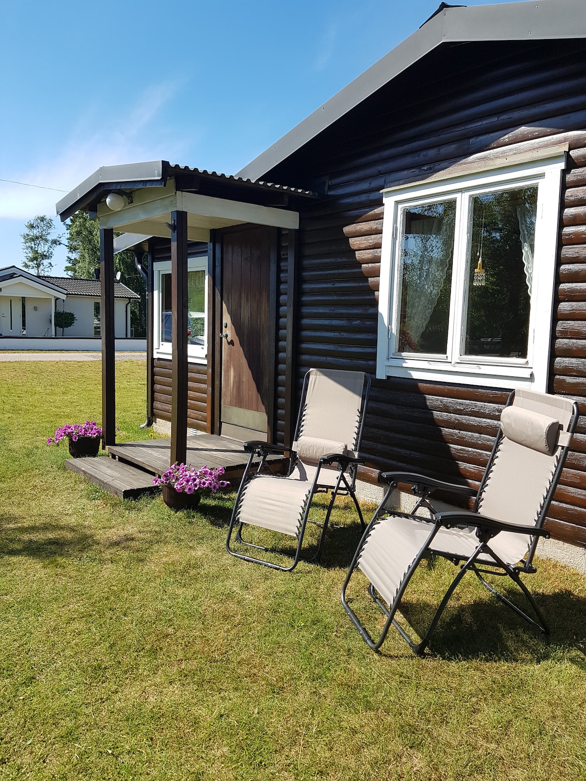 Two comfortable outdoor recliners are positioned on a wooden porch, accompanied by flowerpots containing vibrant purple blooms. The entrance to a cozy cabin is visible, featuring a dark wooden door and large windows that let in natural light.