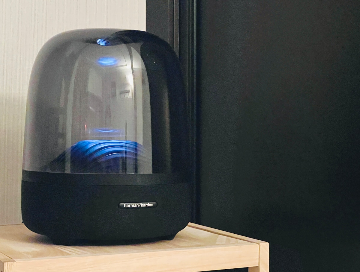 A stylish speaker is positioned on a wooden surface, featuring a modern, rounded shape and a translucent cover. Subtle blue lighting can be seen illuminating the interior, creating a contemporary aesthetic within the space.