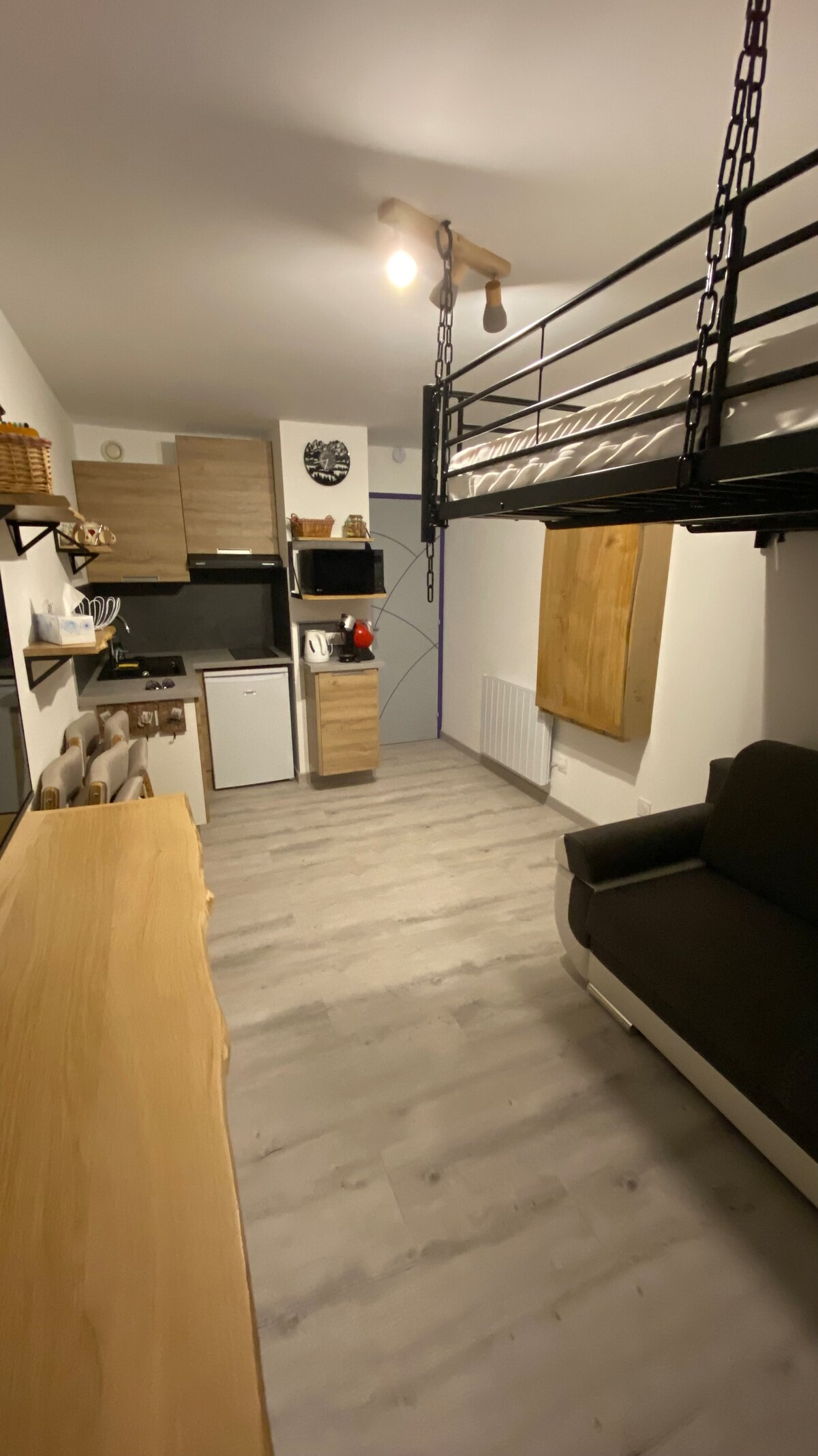 The studio presents an open layout featuring a convertible sofa and a lofted sleeping area with a black metal frame. A kitchenette, equipped with essential appliances, is located along one wall. Light wood accents provide warmth to the space, illuminated by overhead lighting.