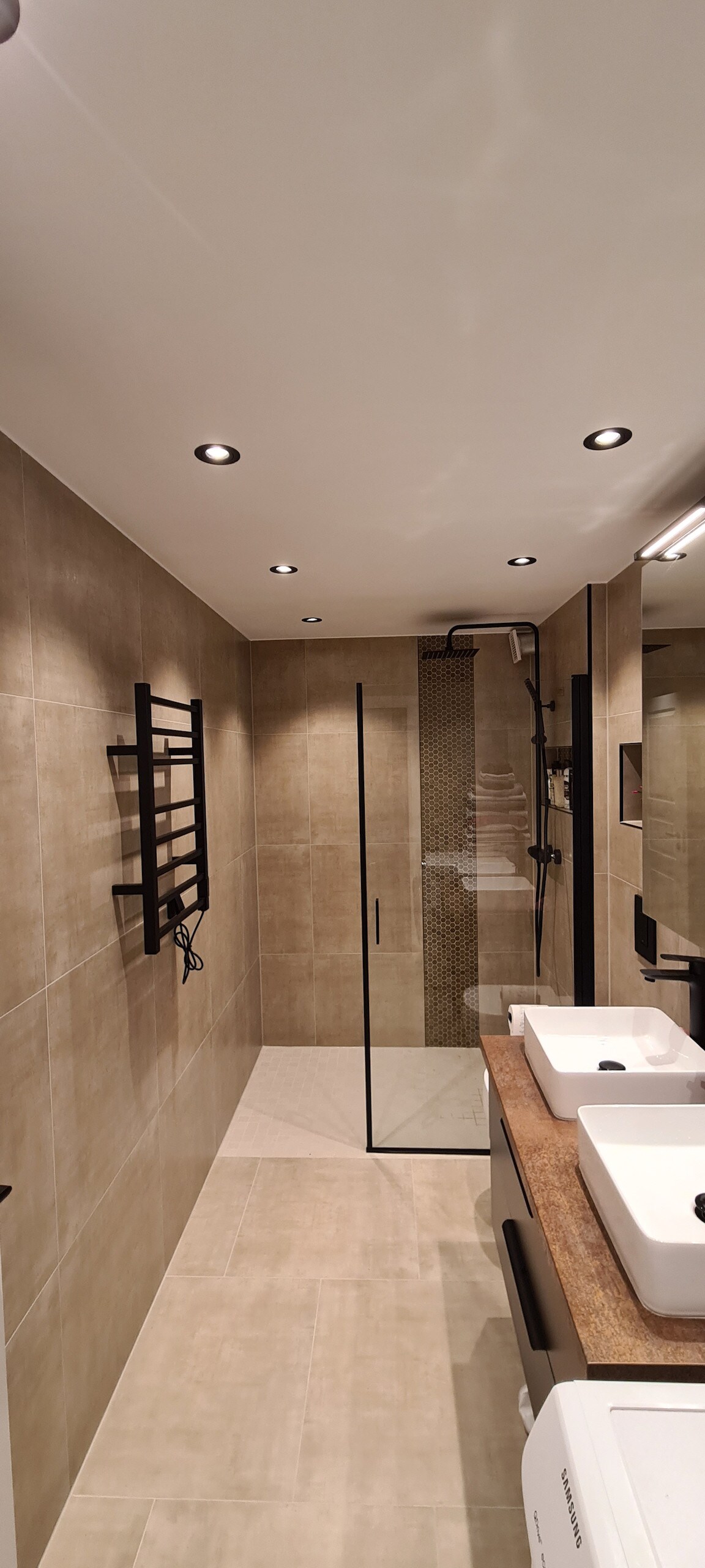 The bathroom features a contemporary design with beige tiled walls and floors. A glass shower enclosure is visible, complemented by a patterned accent wall. Two modern sinks are positioned on a wooden countertop, and wall-mounted fixtures provide illumination.