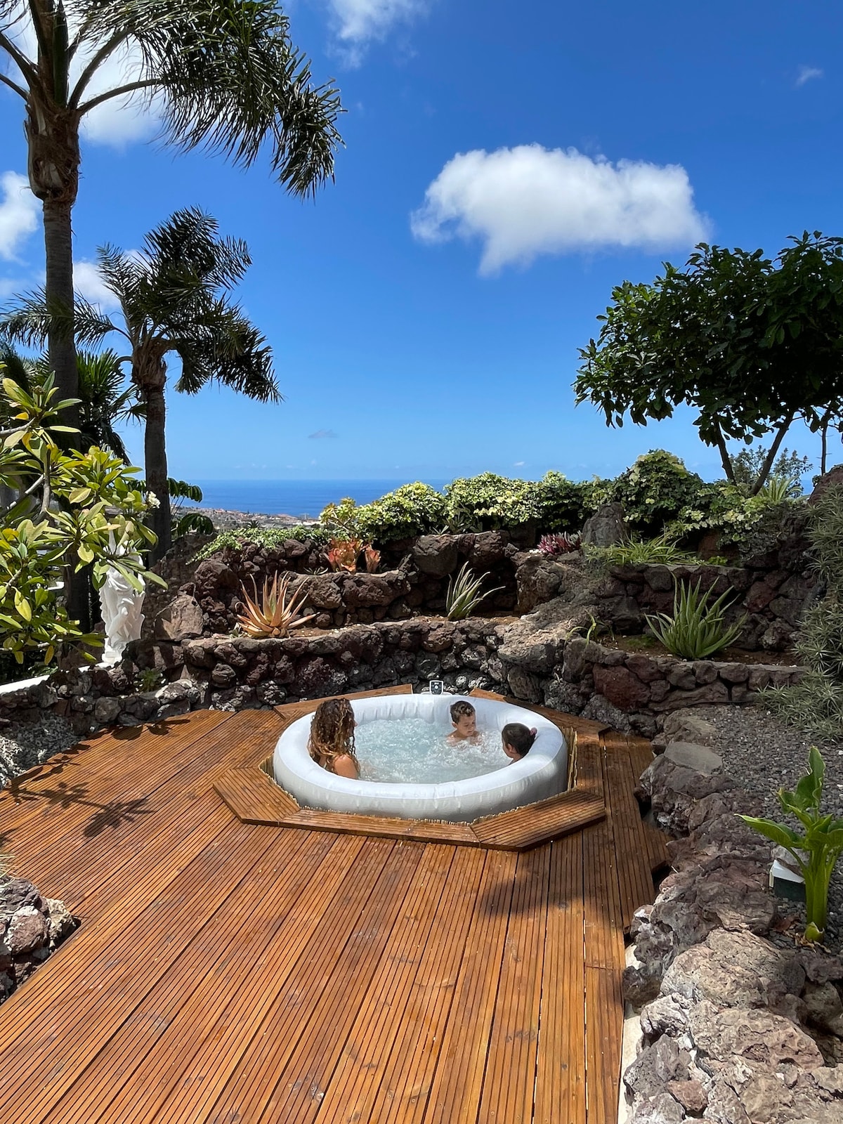 A hot tub is set on a wooden deck, surrounded by a vibrant garden featuring various tropical plants. The blue sky and ocean are visible in the background, complementing the tranquil outdoor atmosphere.