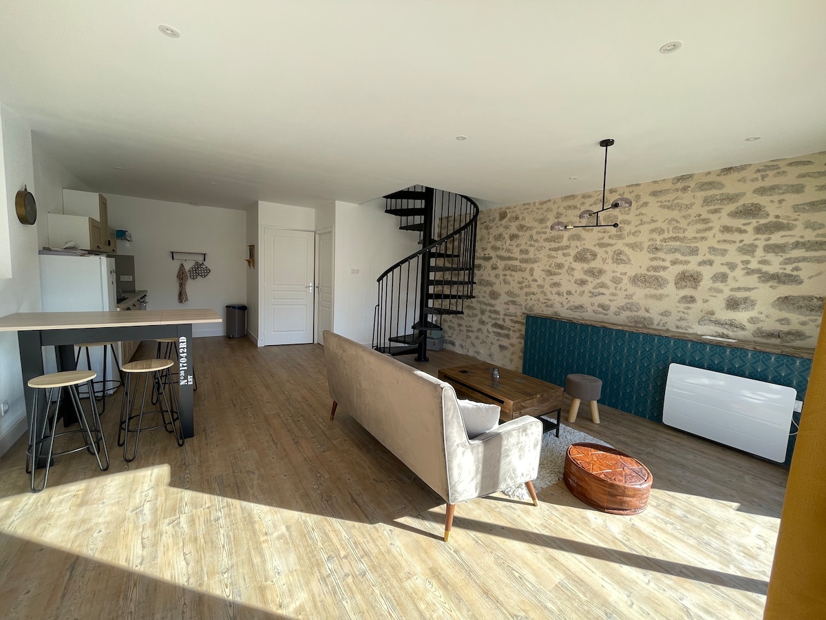 An open living area features a spiral staircase leading to an upper level, with a neutral-toned sofa and wooden coffee table at the center. Exposed stone walls add texture, while natural light floods the space through large windows, creating a bright and airy atmosphere.