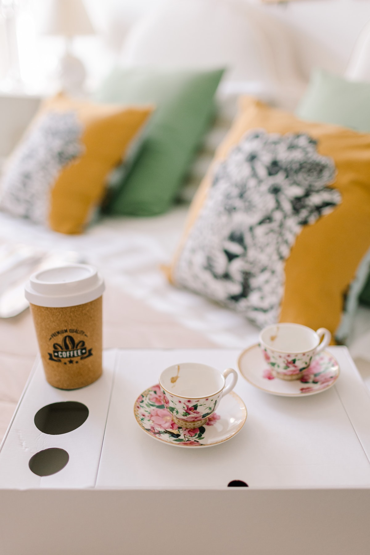 A small white table is set with delicate floral teacups and saucers. A coffee cup with a cardboard sleeve sits next to the teacups. The background features a bed adorned with teal and mustard cushions, creating a cozy setting.