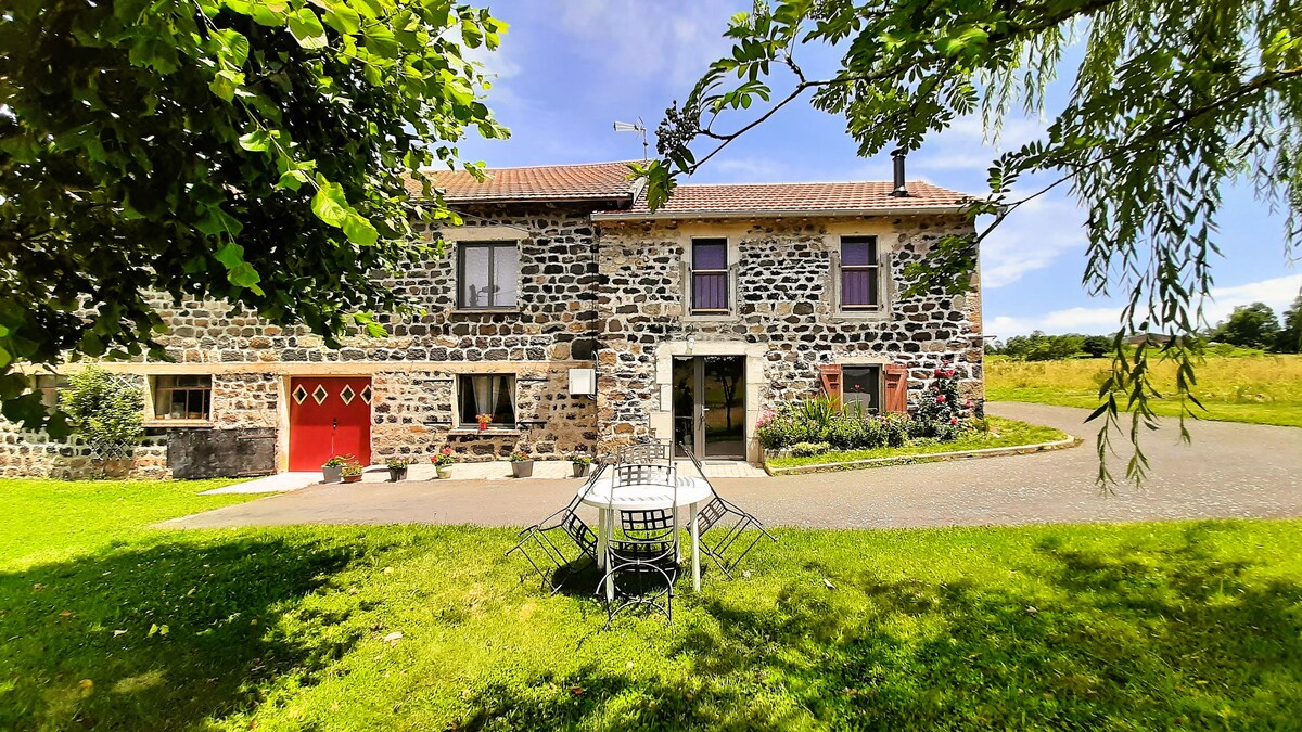 A charming stone farmhouse is set in a green landscape, featuring a well-maintained exterior with red doors and windows. A small outdoor table and chairs are placed on a grassy area, surrounded by colorful flowerbeds. The scene is enhanced by a clear blue sky and gentle sunlight.
