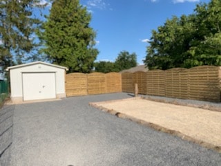 An outdoor area is presented, featuring a gravel surface and a storage shed situated against a backdrop of wooden fencing. Tall trees provide shade, while clear blue skies and scattered clouds indicate a pleasant day.