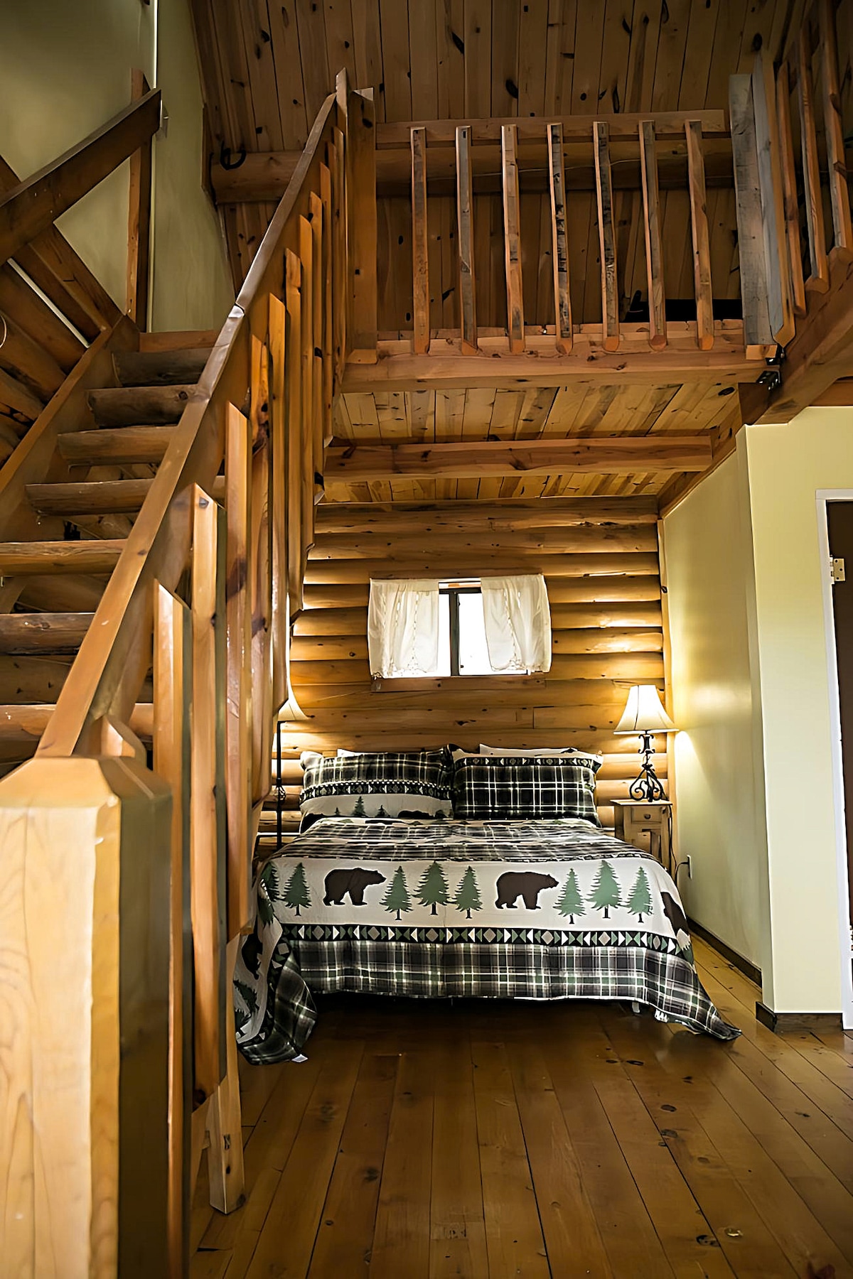 A cozy sleeping area features a log cabin design with warm wooden walls and flooring. A spacious bed is adorned with a patterned blanket depicting bears and trees. A bedside lamp illuminates the area, complemented by a small window allowing natural light to enter.