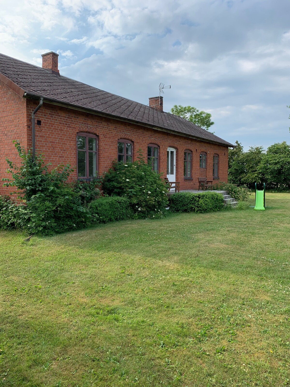 A charming red-brick house is set amidst a lush green lawn, featuring garden hedges, shrubs, and a slide in the foreground. The building showcases large windows and a set of steps leading to a door, inviting exploration of the serene outdoor space.
