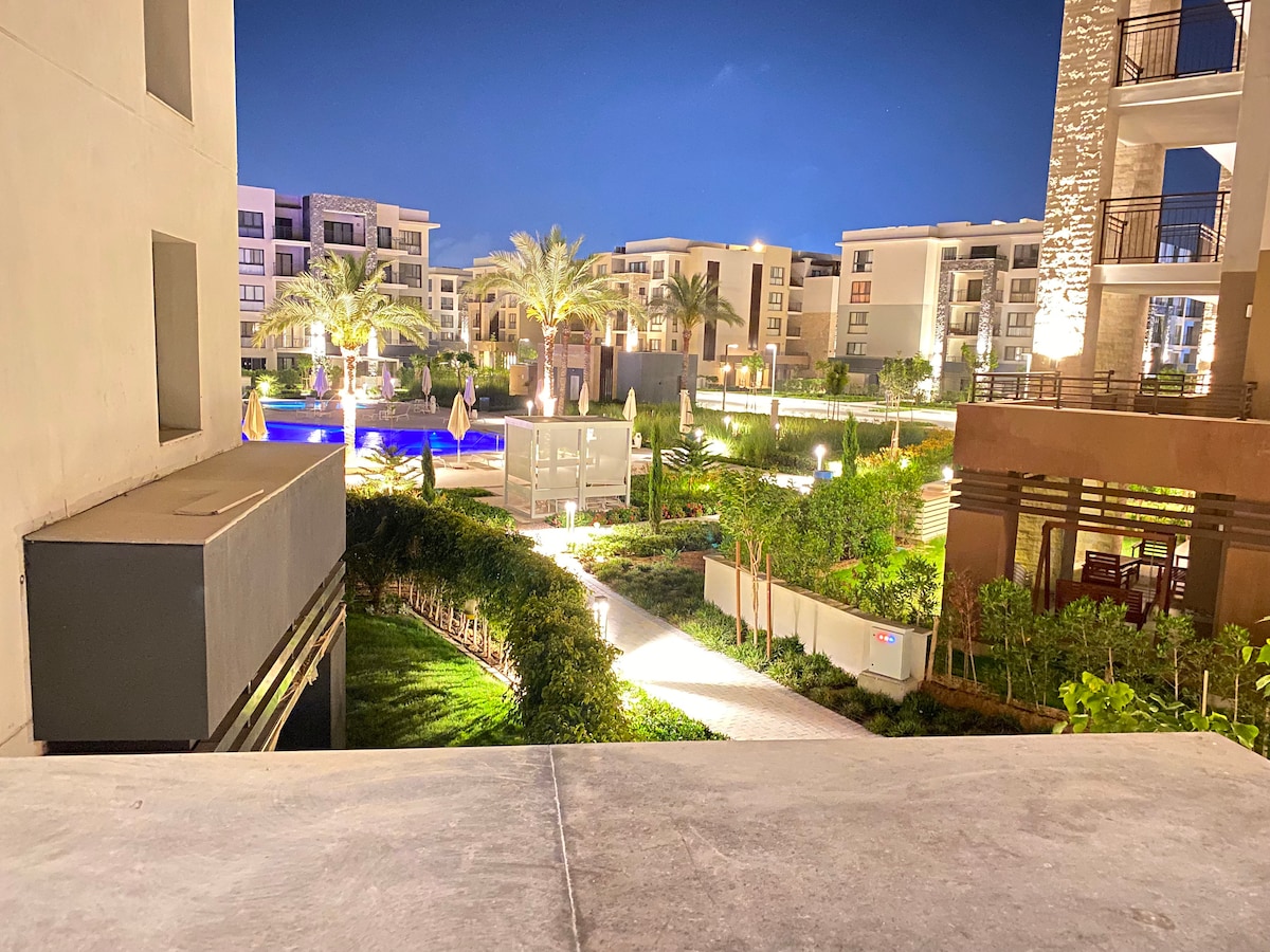 The view from the balcony captures a well-lit outdoor area featuring palm trees and landscaped gardens. A swimming pool is visible in the background, surrounded by comfortable seating. Soft lighting enhances the serene atmosphere, creating an inviting environment for evening relaxation.