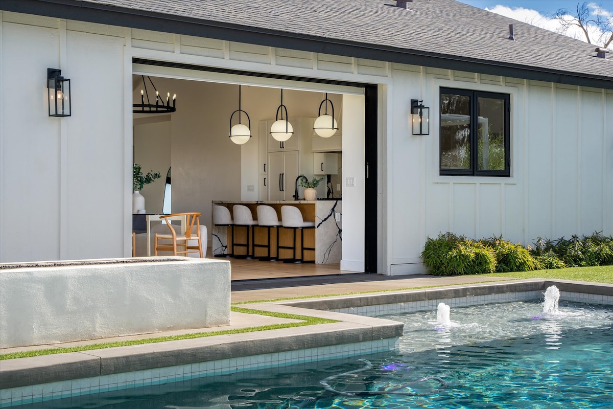 The view showcases a serene pool with gentle water features and a well-maintained lawn. A spacious open kitchen and dining area can be observed through the large pocket sliding door, illuminated by pendant lights, enhancing the connected indoor and outdoor living experience.